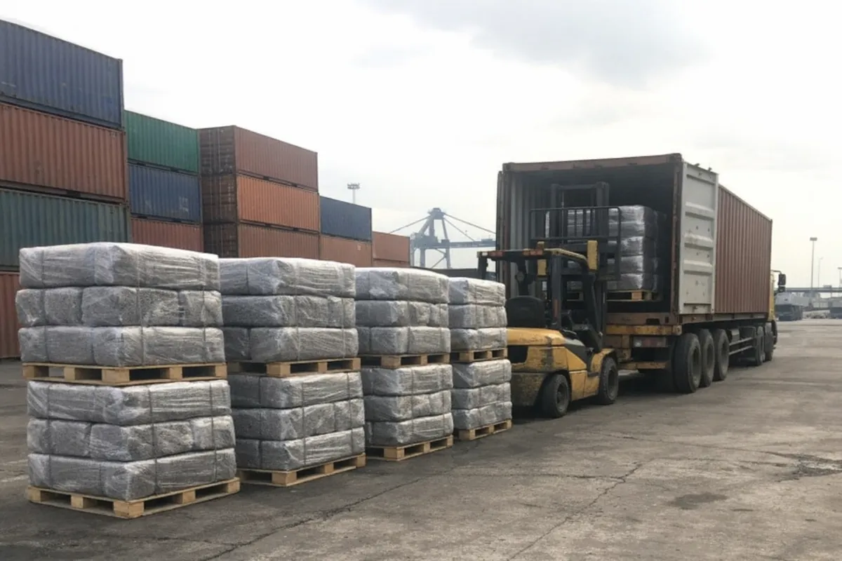 Shrink-wrapped pallets of TSR bales staged beside an open 20-foot container at Belawan port, forklift parked nearby with container yard beyond.